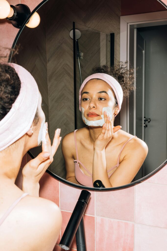 Young woman applying a face mask while looking in the mirror, enjoying her skincare routine.