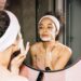 Young woman applying a face mask while looking in the mirror, enjoying her skincare routine.